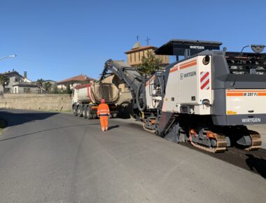 Macchina asfaltatrice e camion su strada, operaio in arancione.