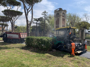 Operai asfaltano strada con macchinari, camion e alberi sullo sfondo.