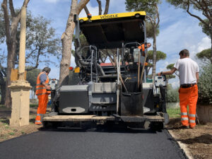 Operai asfaltano strada con macchinario, circondati da alberi.