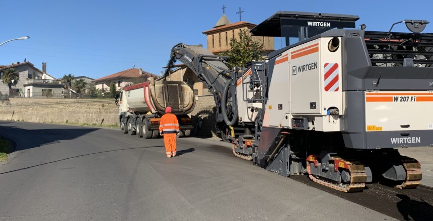 Macchina asfaltatrice e camion su strada, operaio in arancione.